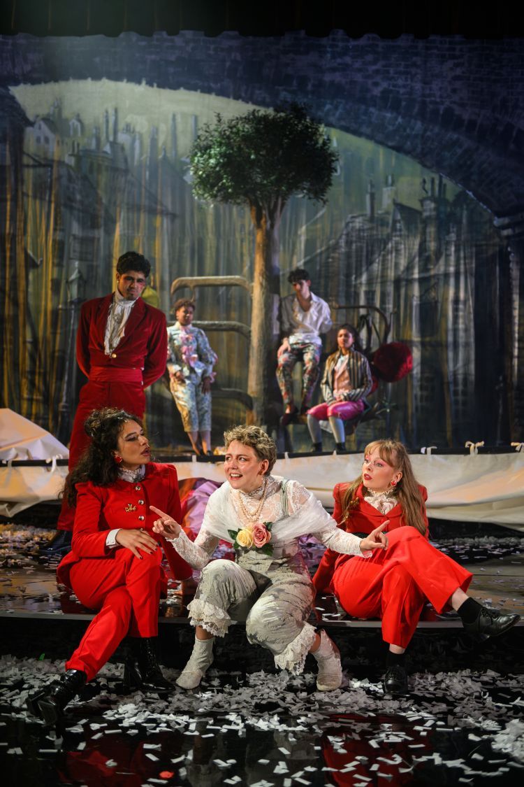 Cast of Orlando on stage, the ensemble are in variations of bright red 70's style suits and the cast members portraying Orlando are in pastel blues, pinks and white renaissance style costumes